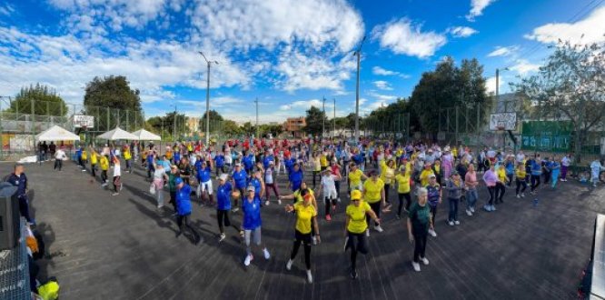 Personas participando en una jornada deportiva al aire libre, reunidas en una cancha asfaltada, en un día soleado. Están distribuidas en grupos con camisetas de diferentes colores (azul, amarillo, rojo y otros), realizando ejercicios o pasos de baile de forma coordinada. 