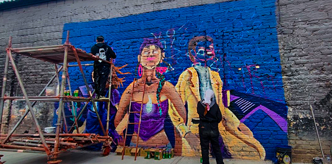 Mural en proceso de pintura sobre una pared de ladrillo en Puente Aranda, donde artistas trabajan en la recuperación y embellecimiento del espacio público cercano a la Cárcel Modelo, reemplazando el deterioro urbano por expresiones artísticas coloridas.