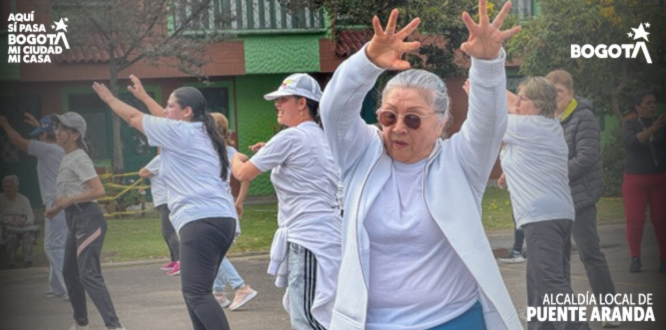 Mujer adulta mayor con gafas oscuras levanta los brazos mientras participa en una clase de ejercicio al aire libre junto a otras personas en una cancha del barrio.