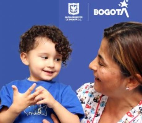 "Mujer sonríe mientras sostiene en brazos a un niño pequeño, ambos mirándose con ternura, sobre un fondo azul con los logos de la Alcaldía Mayor de Bogotá y Bogotá."