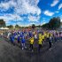 Personas participando en una jornada deportiva al aire libre, reunidas en una cancha asfaltada, en un día soleado. Están distribuidas en grupos con camisetas de diferentes colores (azul, amarillo, rojo y otros), realizando ejercicios o pasos de baile de forma coordinada. 