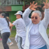 Mujer adulta mayor con gafas oscuras levanta los brazos mientras participa en una clase de ejercicio al aire libre junto a otras personas en una cancha del barrio.