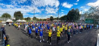 Personas participando en una jornada deportiva al aire libre, reunidas en una cancha asfaltada, en un día soleado. Están distribuidas en grupos con camisetas de diferentes colores (azul, amarillo, rojo y otros), realizando ejercicios o pasos de baile de forma coordinada. 