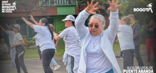 Mujer adulta mayor con gafas oscuras levanta los brazos mientras participa en una clase de ejercicio al aire libre junto a otras personas en una cancha del barrio.
