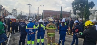 Funcionarios y equipos de emergencia participan en el Simulacro Distrital 2025 en Puente Aranda. En la imagen, se observa a personal de Bomberos Bogotá, brigadistas industriales y trabajadores con cascos y uniformes de seguridad recibiendo instrucciones en una zona industrial. El ejercicio demuestra la organización y el compromiso de la localidad con la prevención y la gestión del riesgo.