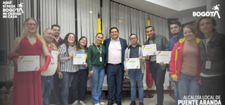 “Grupo de personas posando en un salón de la Alcaldía Local de Puente Aranda luego del acto de posesión del Consejo Local LGBTI 2026–2027. Varias de ellas sostienen certificados en las manos. En el centro se encuentra un funcionario de la Alcaldía acompañado por integrantes electos del Consejo. Al fondo se observan las banderas de Bogotá y de Colombia, y en las esquinas superiores aparecen los logos institucionales de ‘Bogotá’ y de la Alcaldía Local de Puente Aranda.”